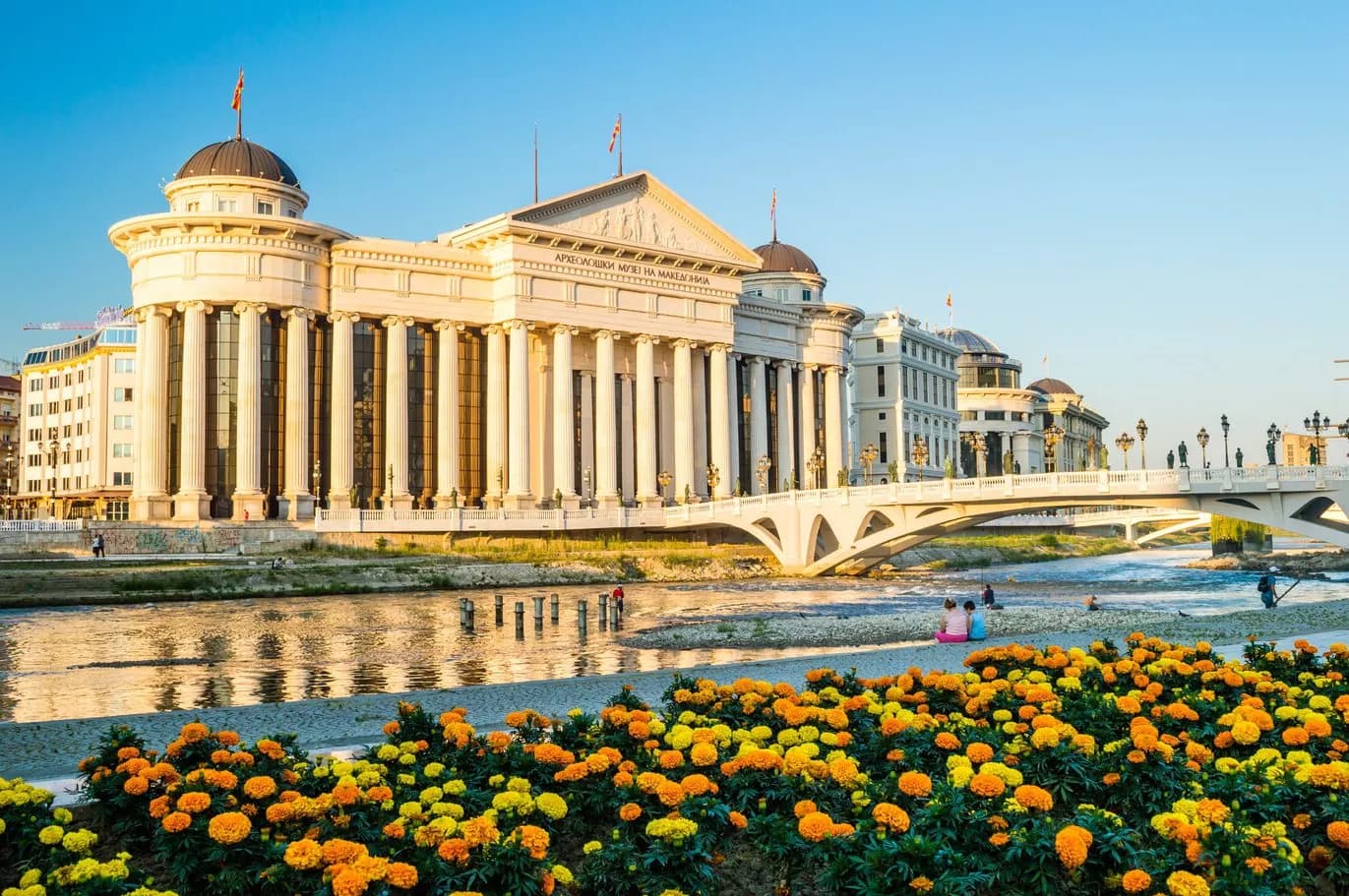Neoclassical museum, bridge, and river in Skopje with bright orange and yellow flowers in the foreground.