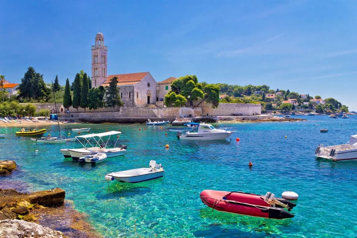 Boats anchored in turquoise water near a stone church with a bell tower on Hvar island.