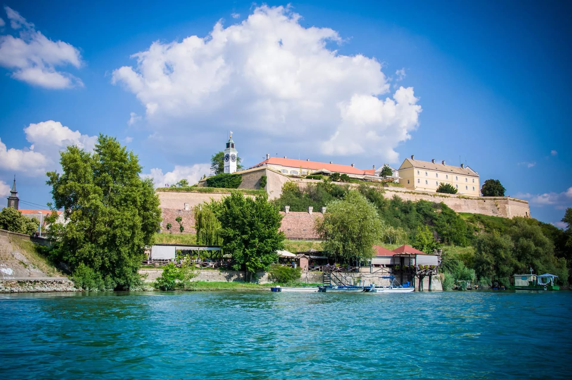 Petrovaradin Fortress on hill above river with boats docked below on sunny day.