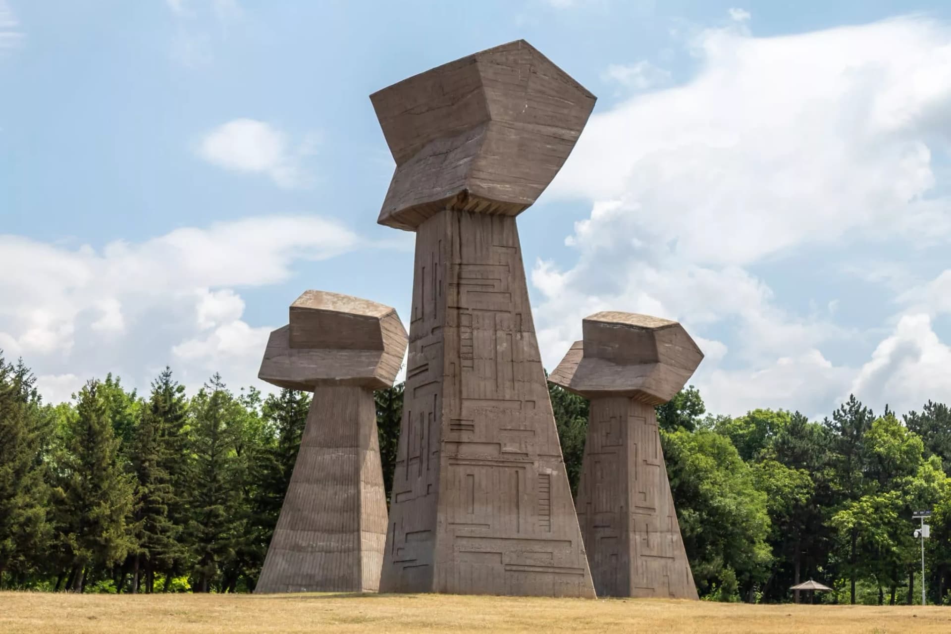 Three Brutalist concrete monuments on dry grass field with green trees and cloudy sky