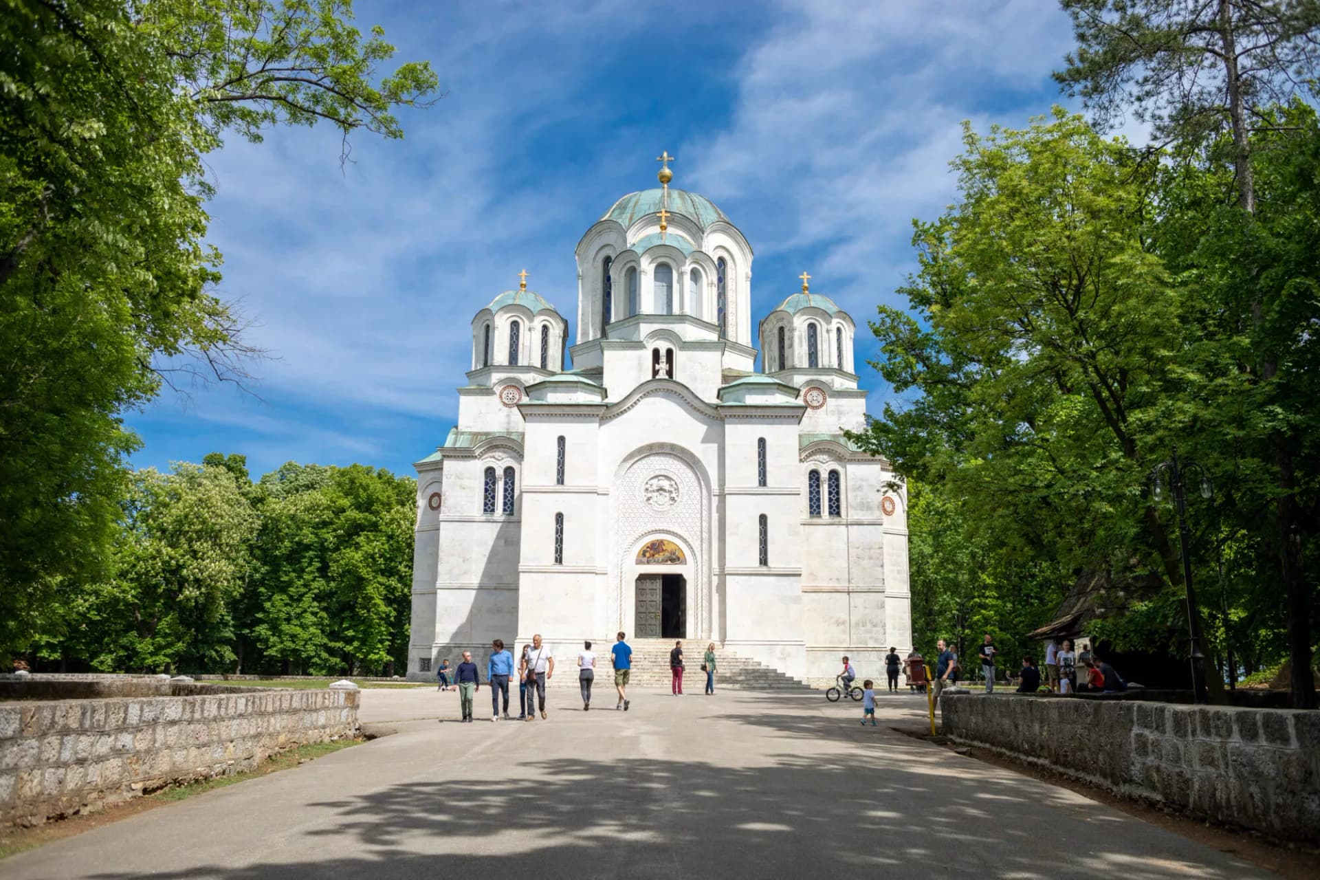White Orthodox church with teal domes surrounded by lush green trees under a blue sky in Oplenac.