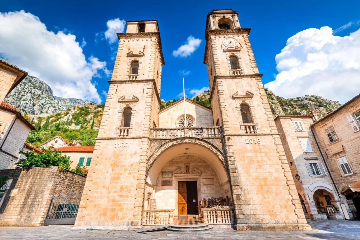 St. Tryphon Cathedral facade with twin bell towers in Kotor, Montenegro, against mountains.