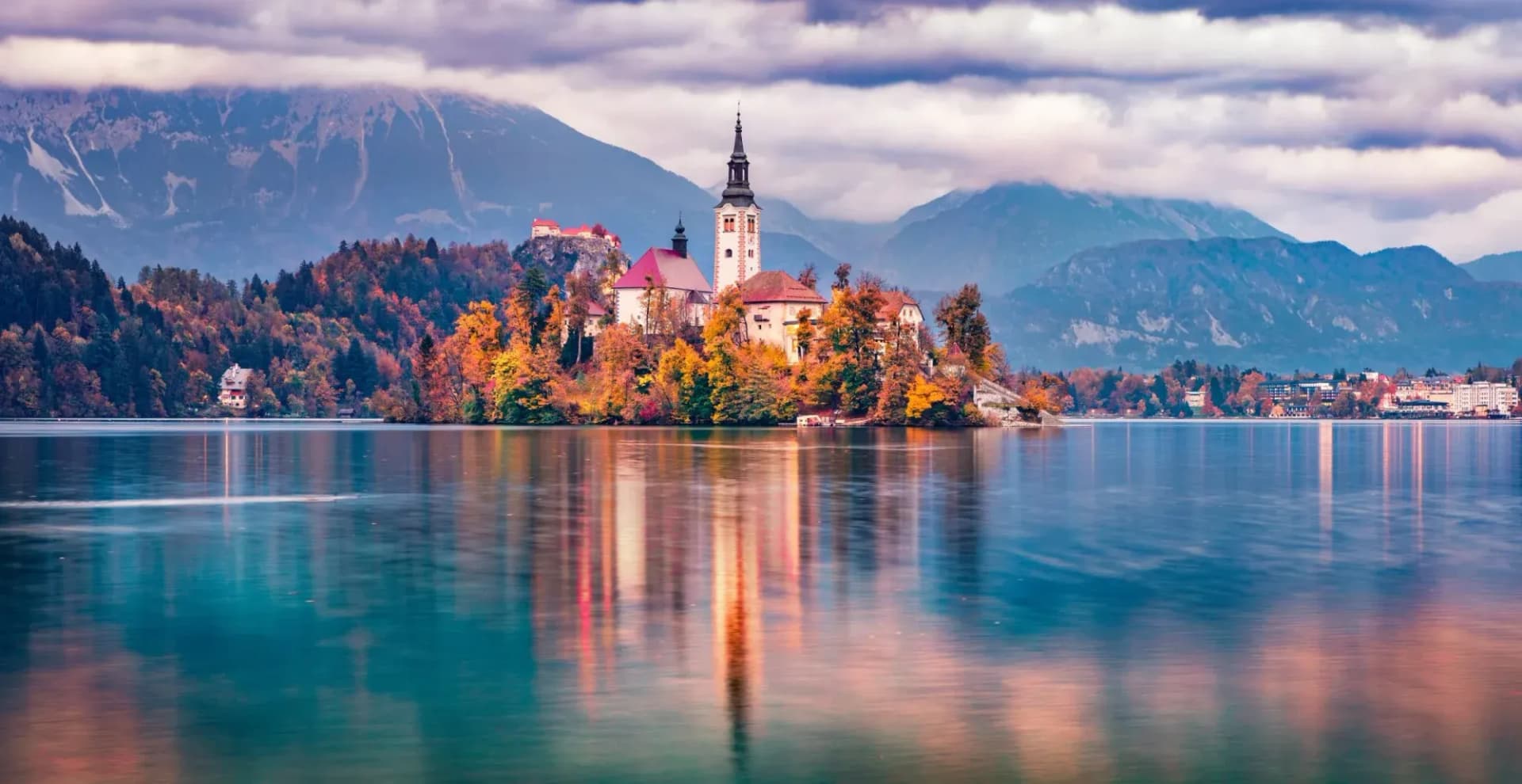 Lake Bled island church with autumn trees reflected in the calm lake and mountains.
