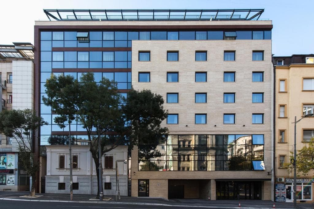 Modern hotel building with glass facade next to older buildings and street trees.