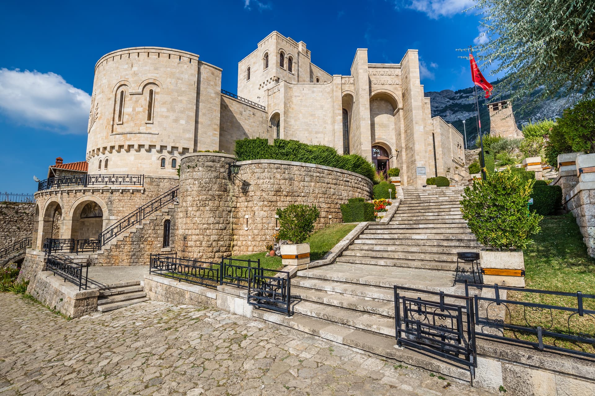 Stone fortress entrance with large stone steps, manicured bushes, and a red flag against a blue sky.