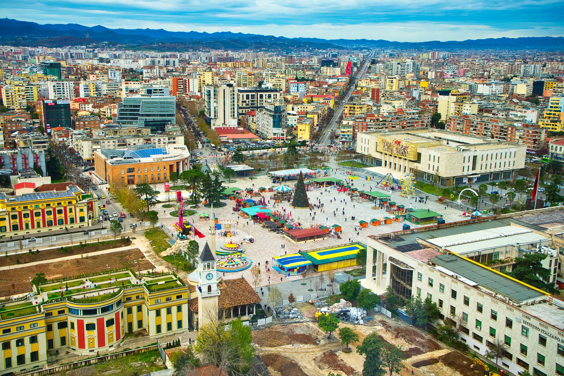 Aerial view of city square with Christmas market, rides, and clock tower against mountain backdrop.