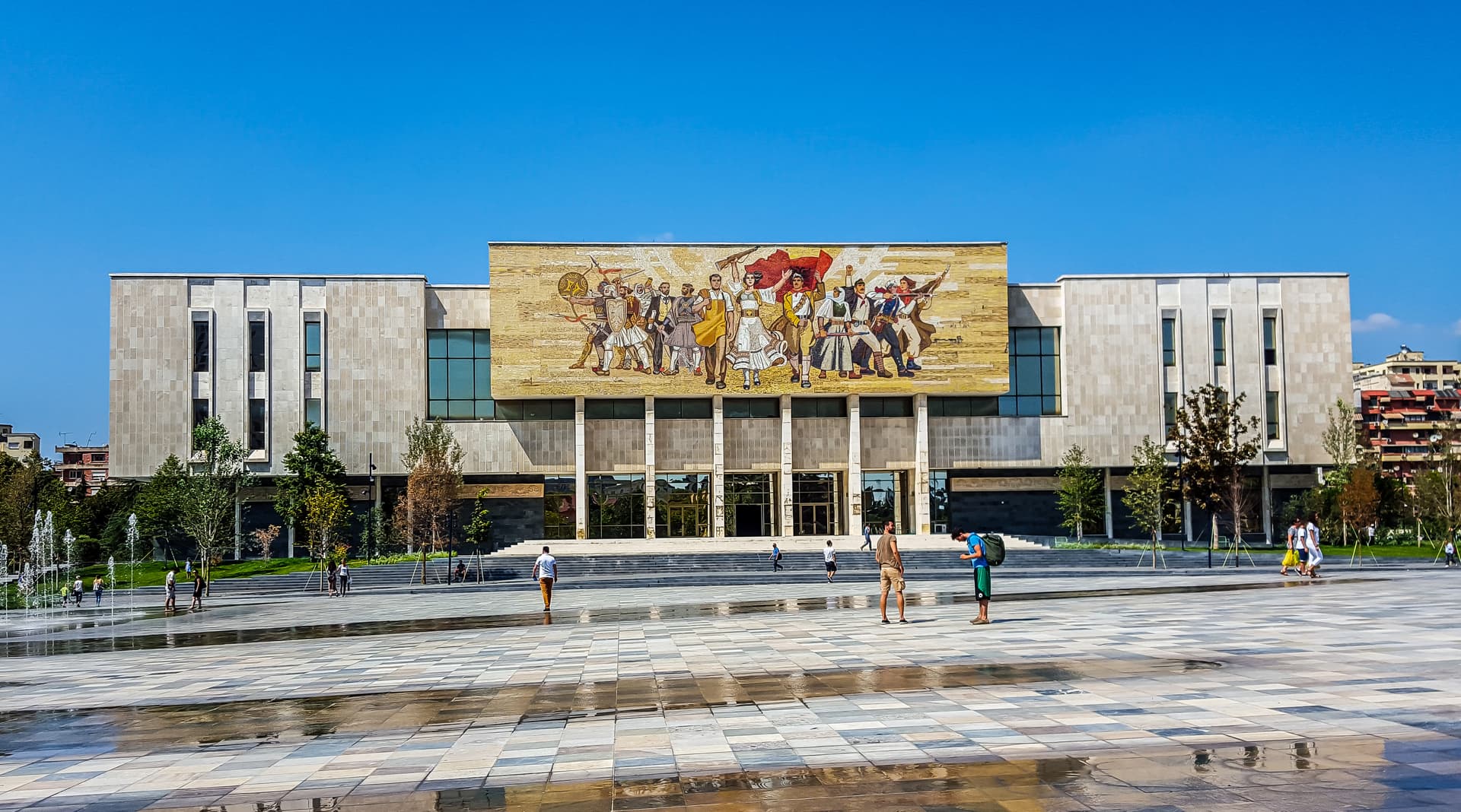 National Historical Museum in Tirana with large mosaic facade and people near plaza fountains.
