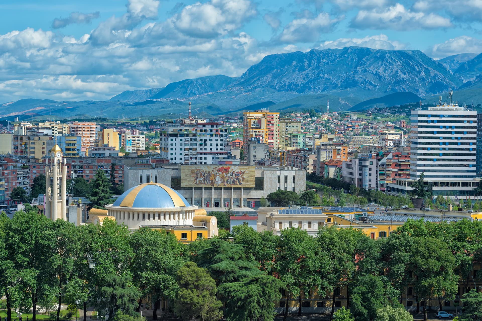 Cityscape of Tirana with colorful buildings, a domed structure, and mountains under a cloudy blue sky.