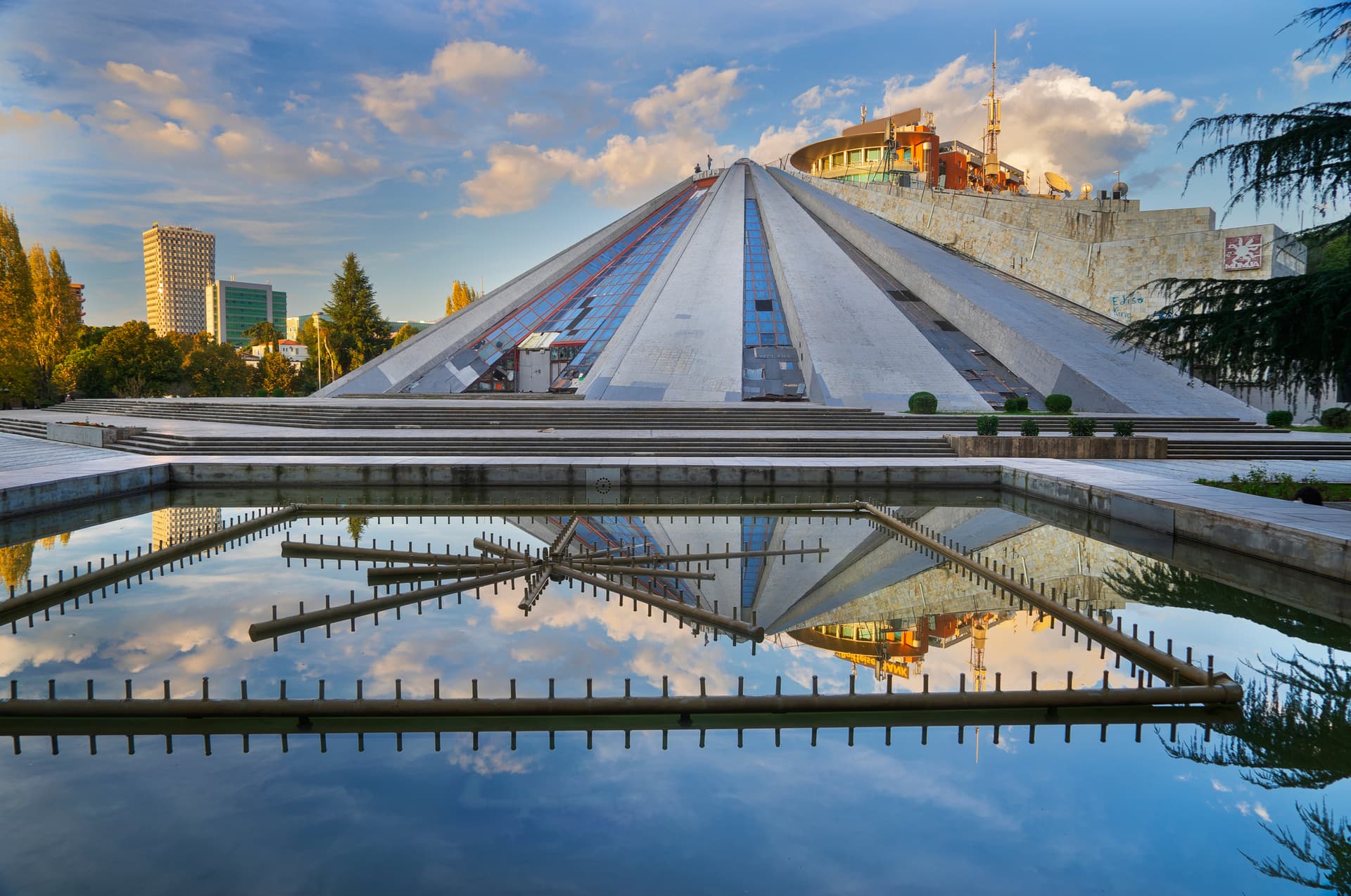 Tirana Pyramid structure reflected in a pool with fountain elements under a cloudy blue sky.