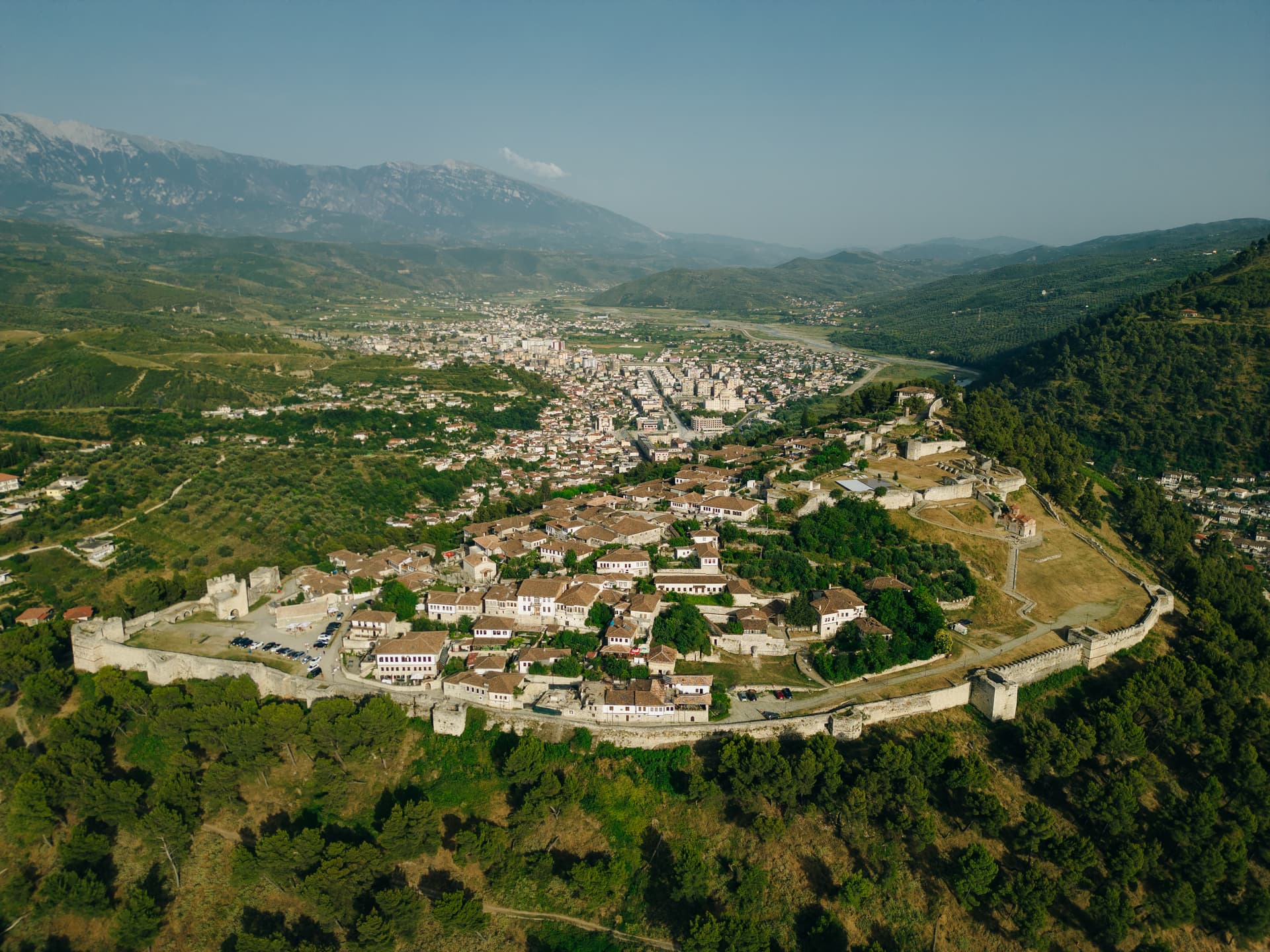 Aerial view of Berat Castle fortress walls overlooking historic Ottoman houses and modern city below mountains.