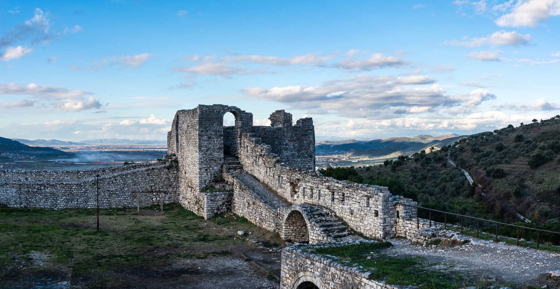 Ruins of Berat Castle stone walls overlooking a valley and distant mountains under a cloudy blue sky.