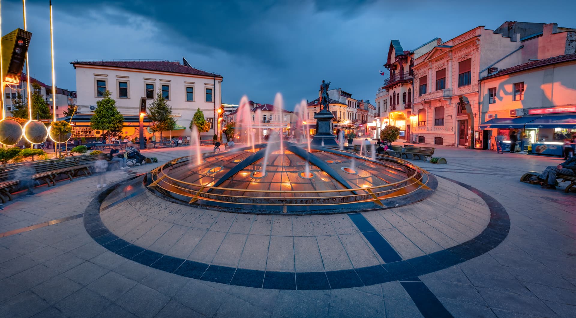 Illuminated fountain in Bitola square at dusk with surrounding historic buildings and people.