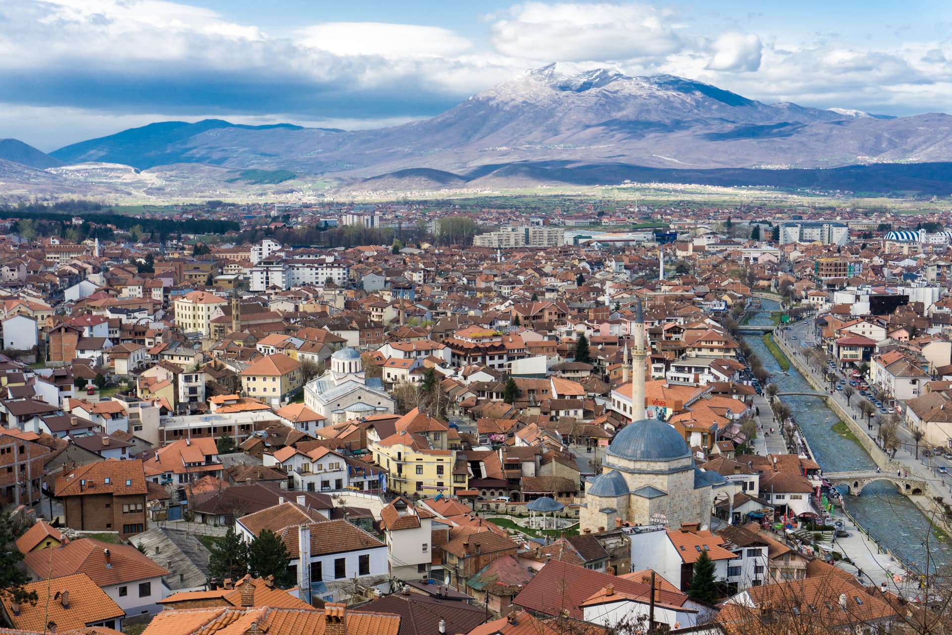 Cityscape of Prizren with terracotta roofs, a river, and snow-capped mountains under a cloudy sky.