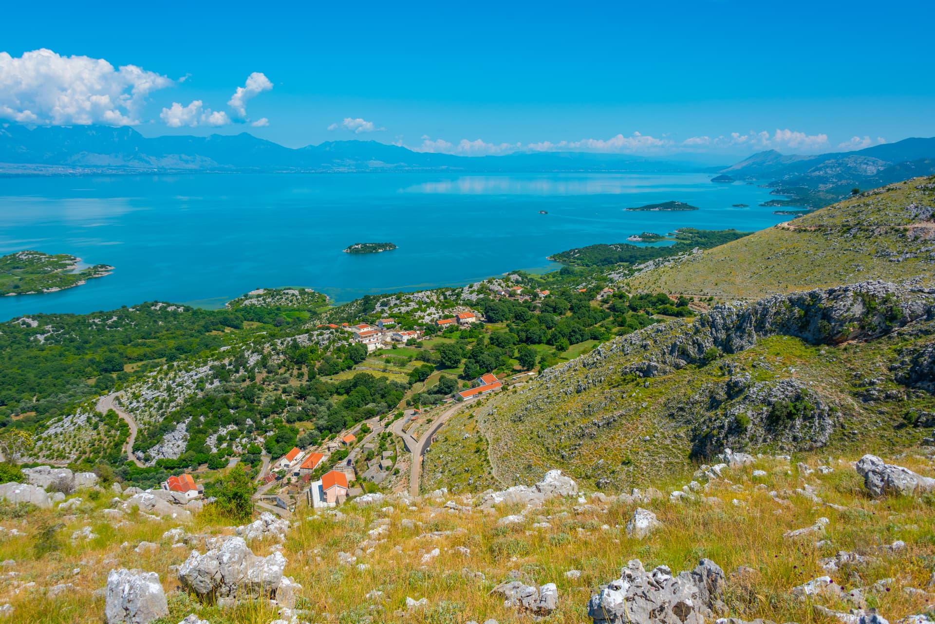 View over Skadar Lake with small village, green hills, and distant mountains under blue sky.