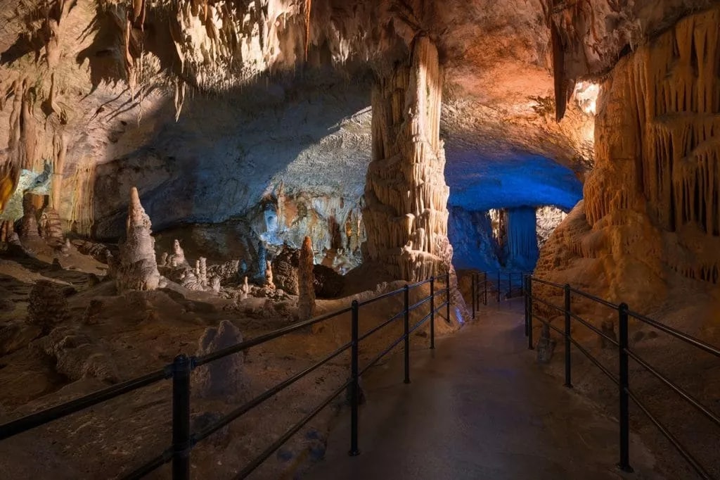 Postojna Cave interior with illuminated stalactites, stalagmites, and a paved pathway with railings.