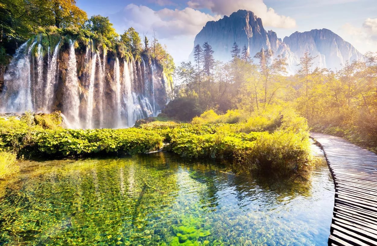 Plitvice Lakes boardwalk over clear water next to waterfalls with a mountain backdrop.