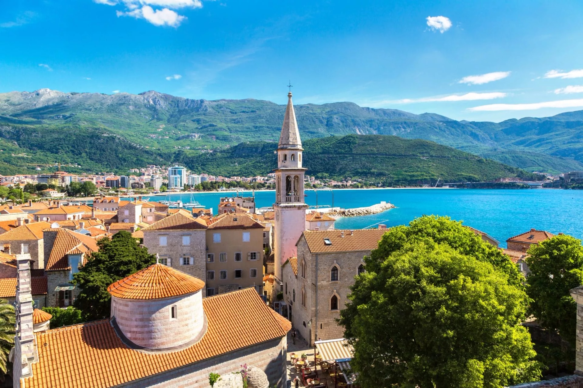 Rooftops of Budva old town with church tower, turquoise sea, and green mountains.