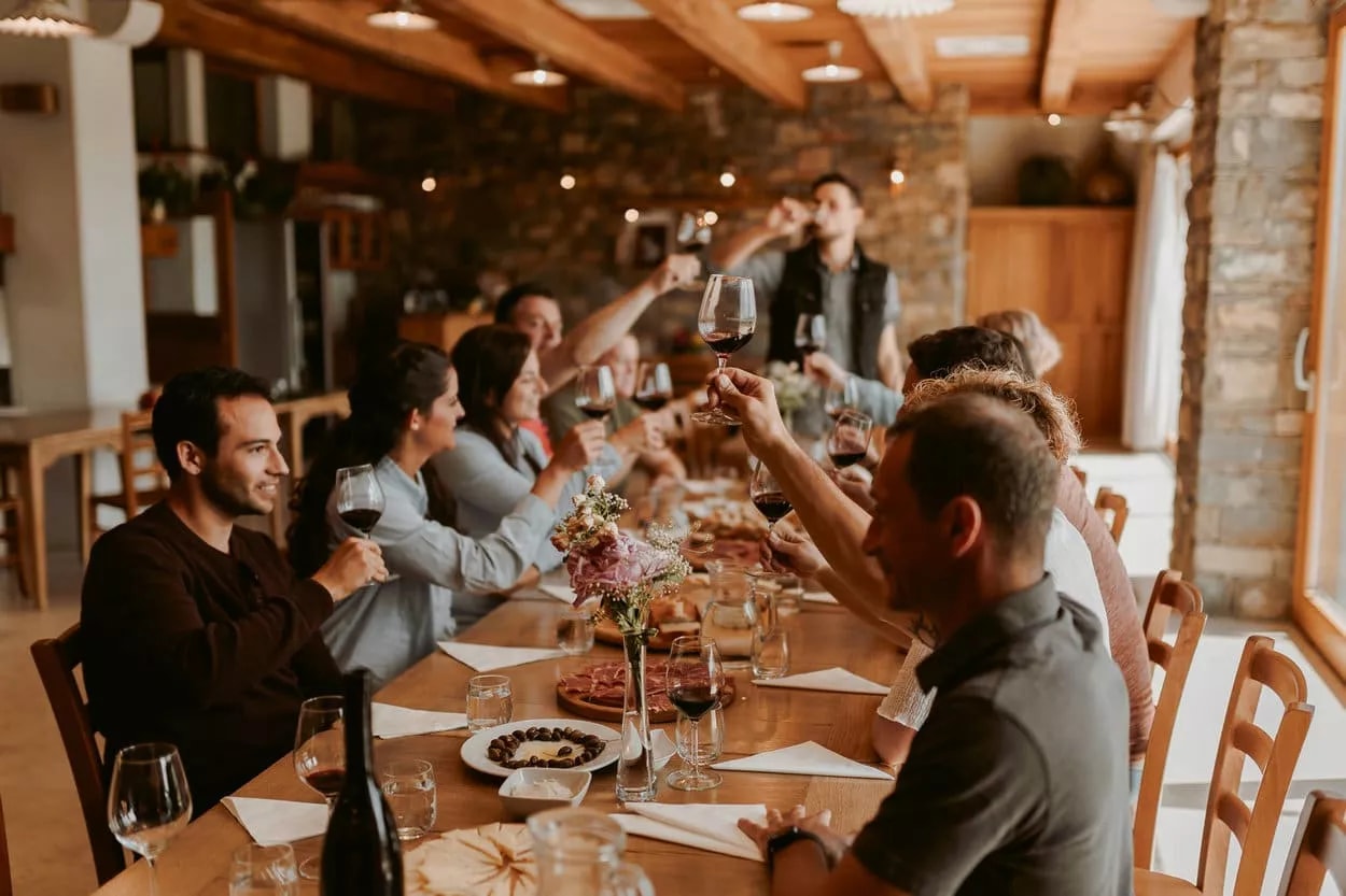 Group toasting with red wine glasses around a wooden table with snacks indoors.