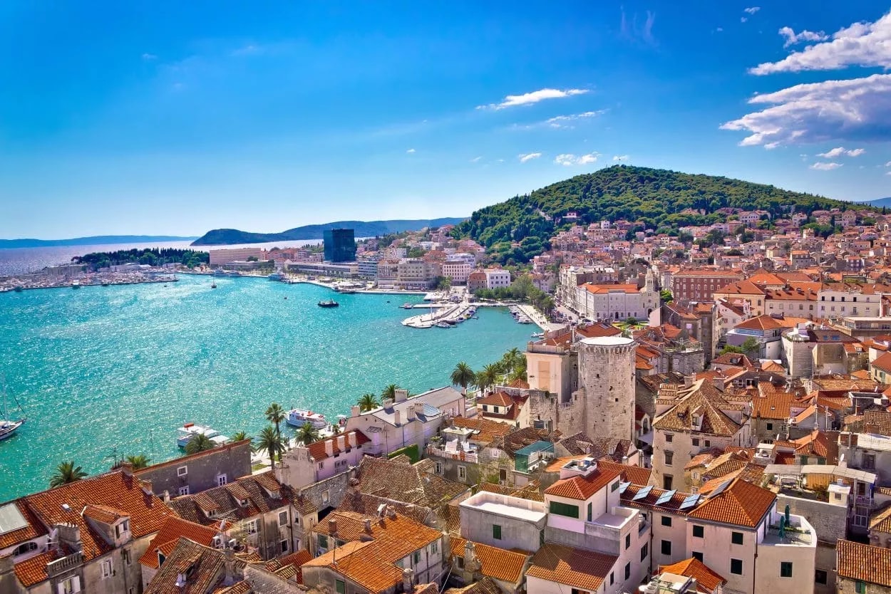 Coastal city with terracotta roofs, turquoise water, and green hill under bright blue sky