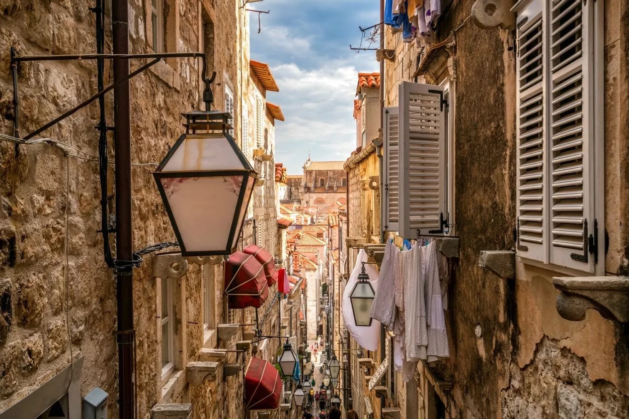 Narrow cobblestone street in Dubrovnik with stone buildings, lanterns, and laundry hanging outside.