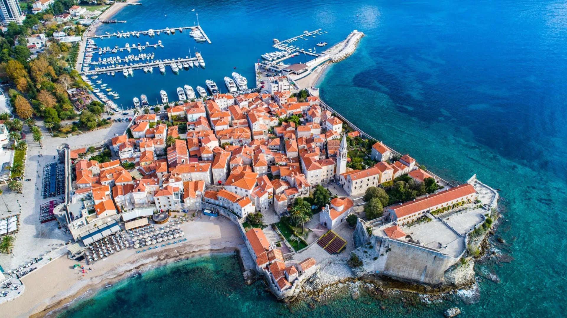 Aerial view of Budva Old Town peninsula with terracotta roofs, harbor, and turquoise Adriatic Sea.