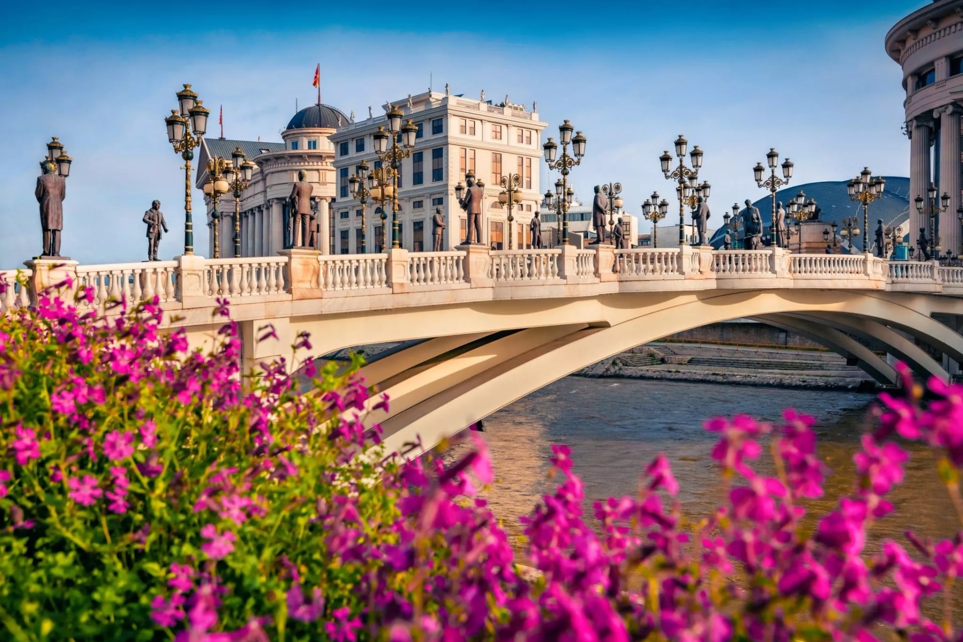 Bridge with statues and ornate lamps over river, foreground of pink flowers, Skopje.