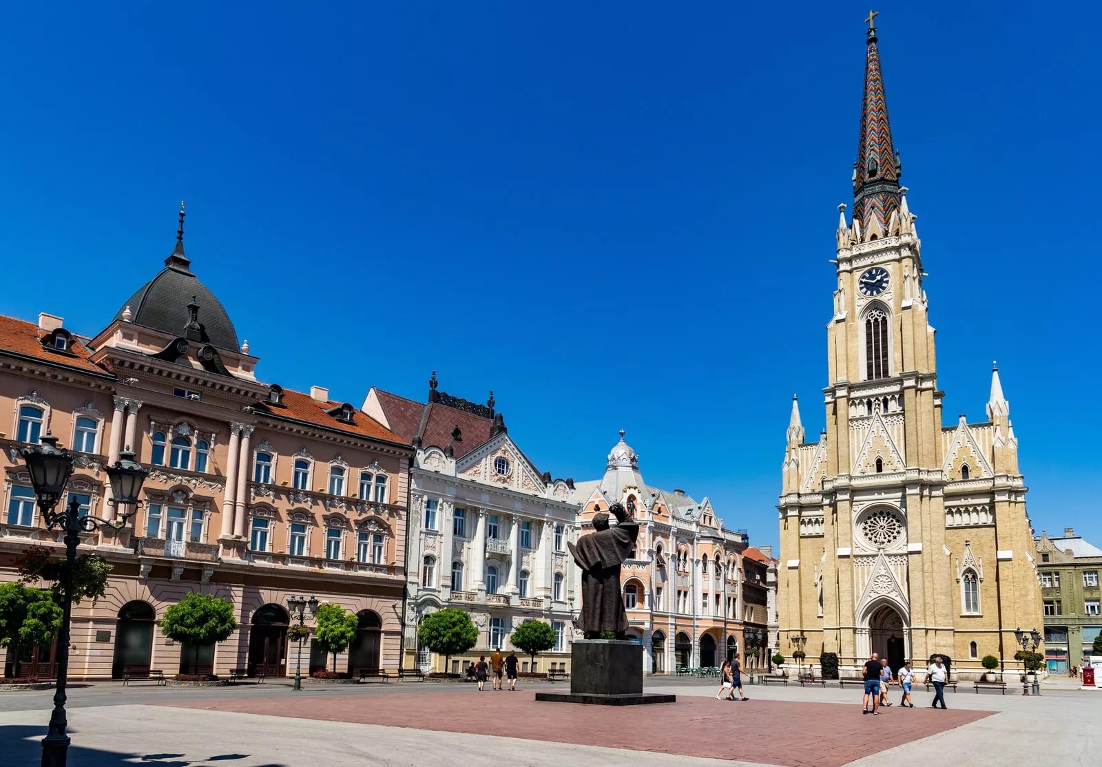 Name of Jesus Cathedral and historic buildings on main square in Novi Sad under clear blue sky.