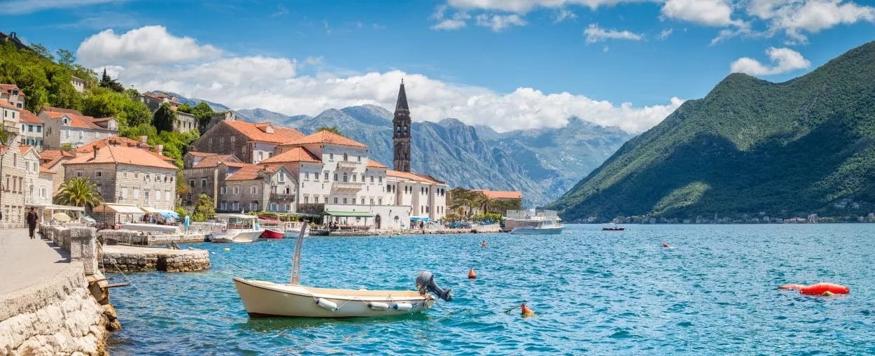 Small boat on blue water near historic stone buildings and green mountains in Kotor Bay, Montenegro.