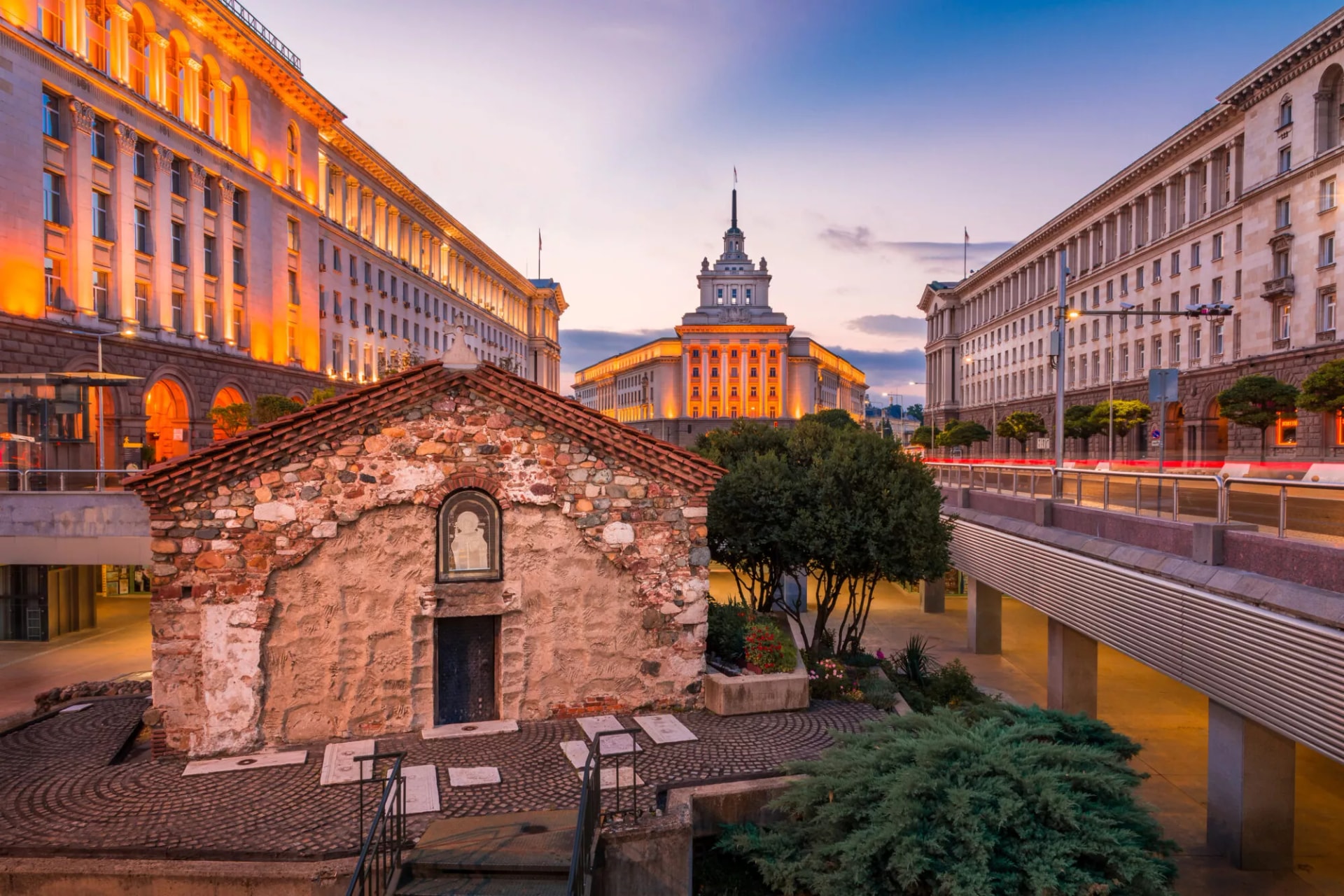 Ancient stone church in Sofia, Bulgaria, framed by illuminated government buildings at dusk.