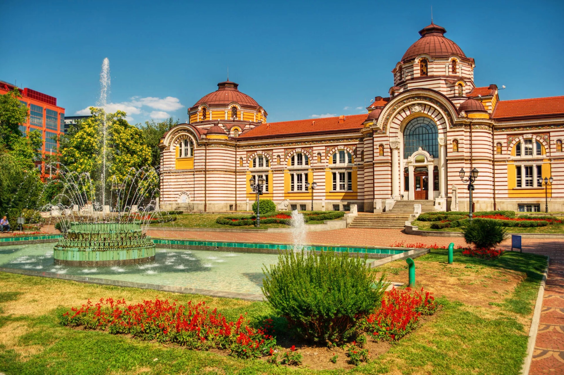 Historic building with striped facade and domes next to a tiered fountain in a park.