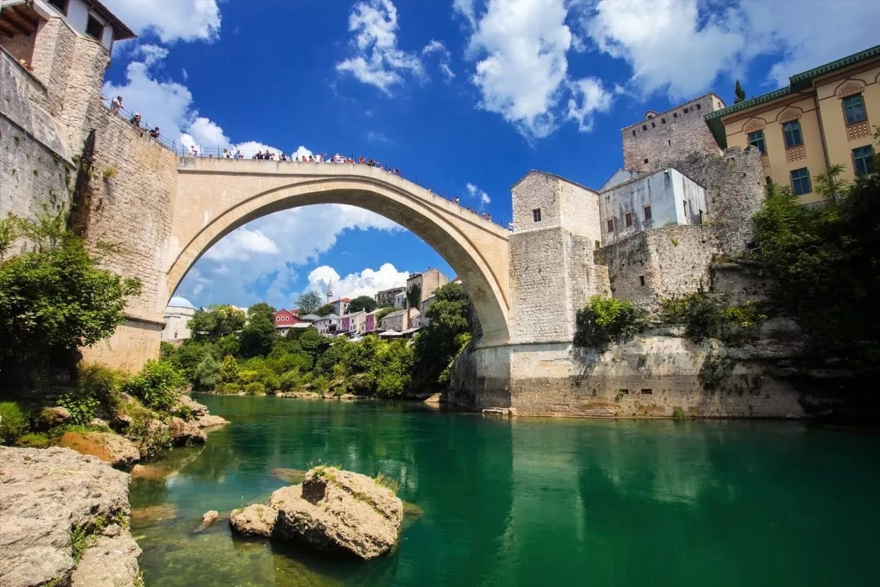 Stari Most bridge over emerald green river with historic stone buildings under blue sky in Mostar.
