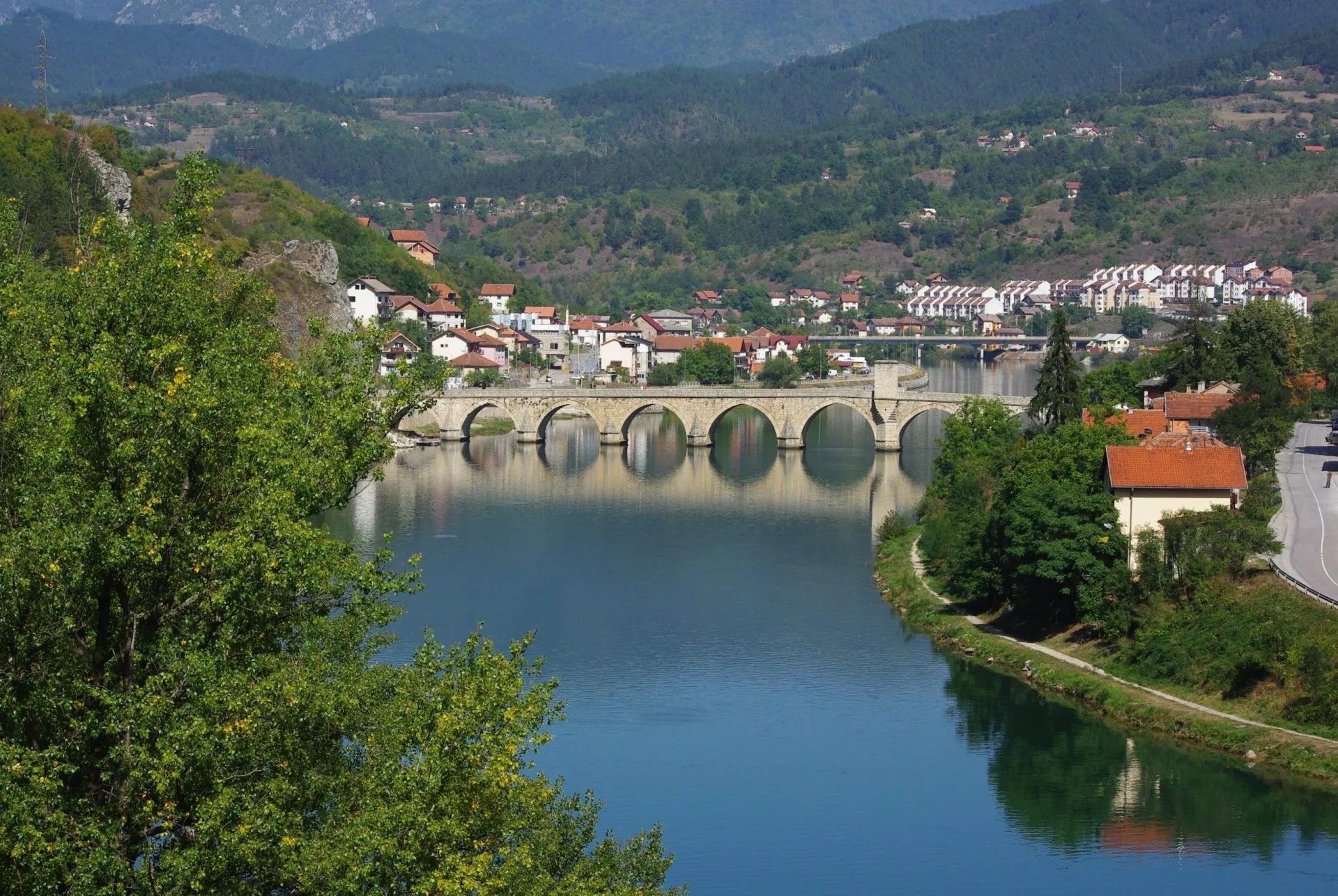Stone arch bridge over river with town and green hills in background, Visegrad.