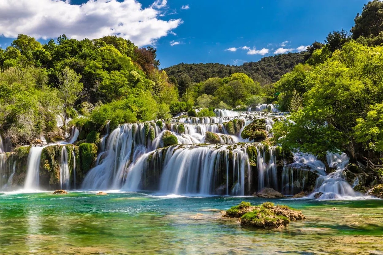 Plitvice waterfalls cascading over mossy travertine barriers into clear turquoise pools under a blue sky.