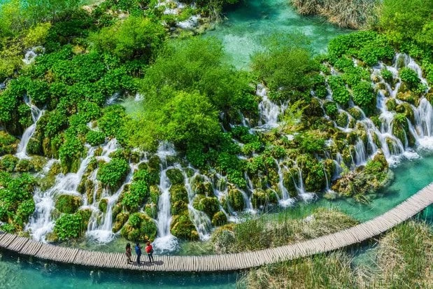 Boardwalk through Plitvice in Croatia with people walking over turquoise water and lush green waterfalls.