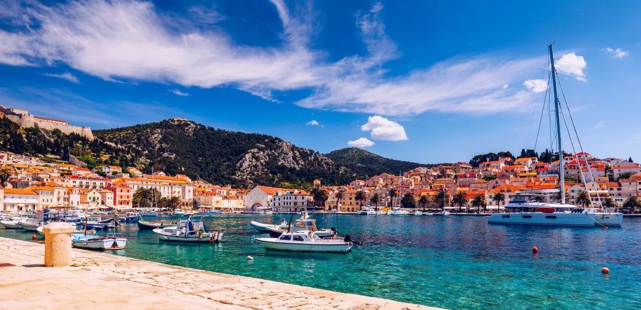 Boats moored in turquoise harbor water with Hvar town and green hills under blue sky.