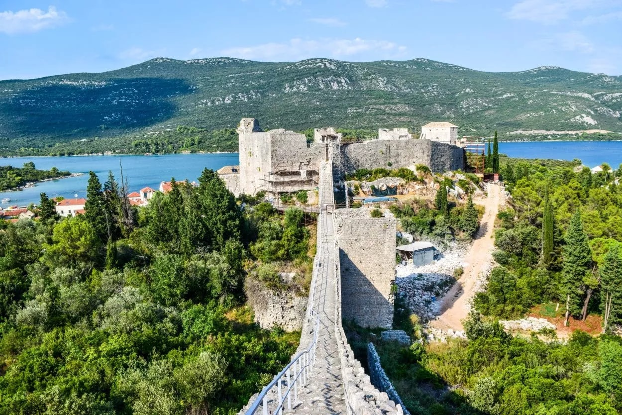 Stone walls and ramparts leading to a medieval fortress overlooking a blue bay and green mountains.