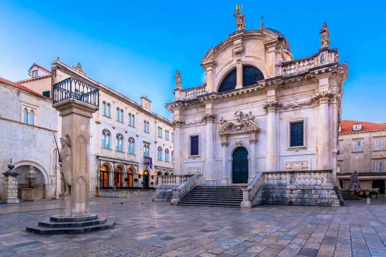 Baroque church facade, stone column with statue, and historic buildings in Old Square in Dubrovnik.