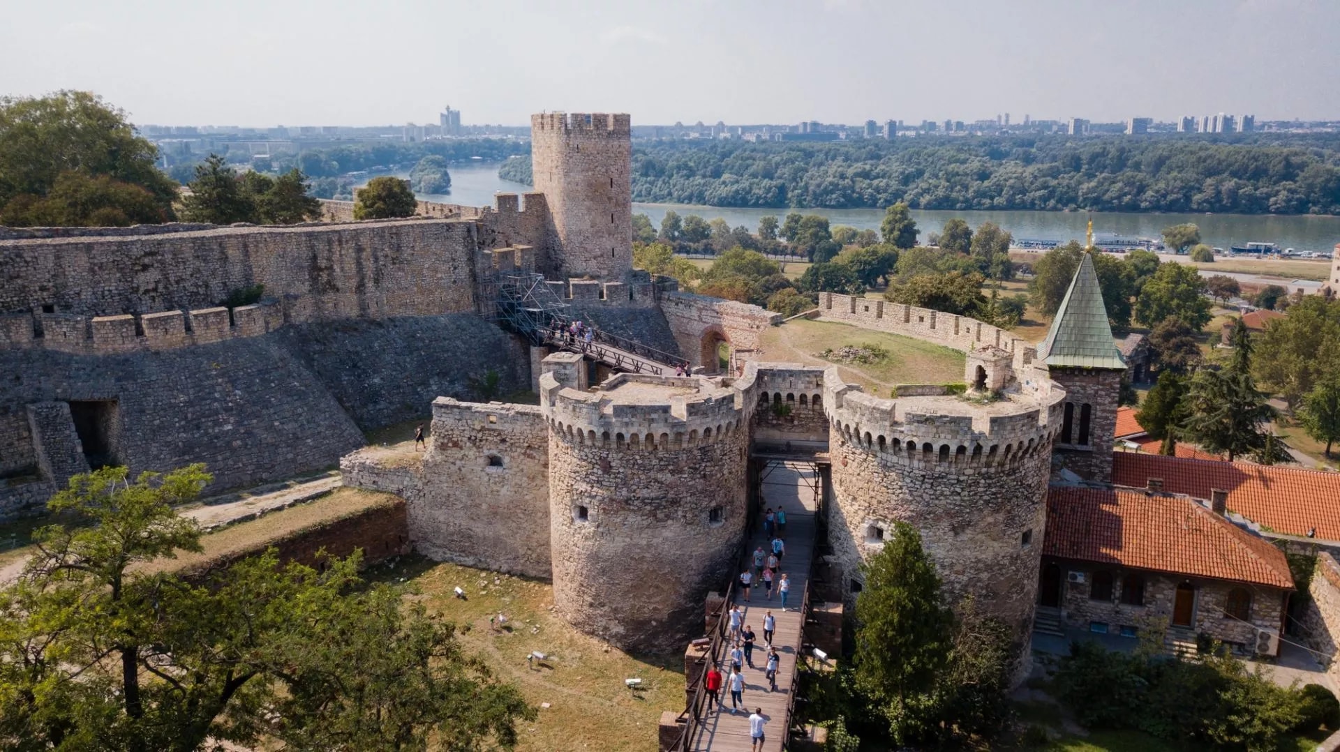 Kalemegdan Fortress stone walls and towers with visitors crossing a wooden bridge, overlooking the river and Belgrade skyl...