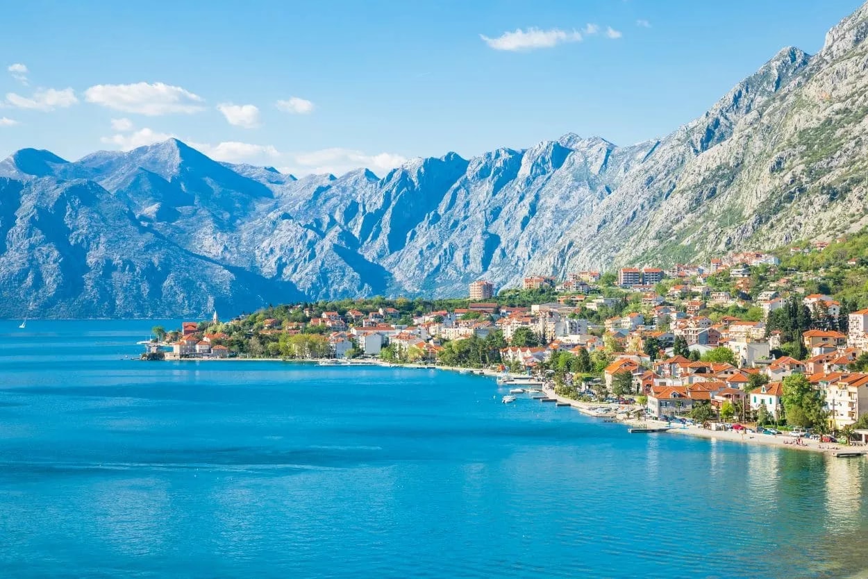 Coastal town with red-roofed buildings nestled against steep mountainsides above bright blue water of the Bay of Kotor.