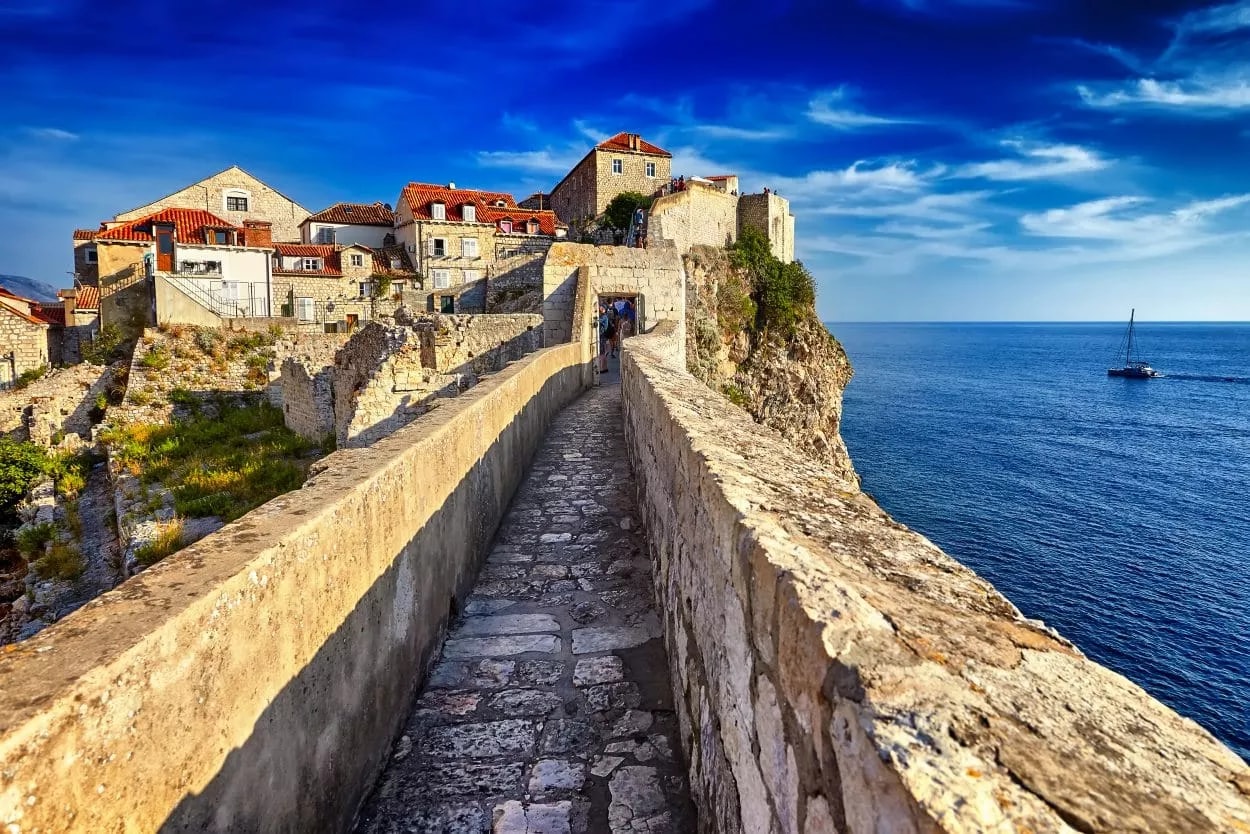 Stone walkway on Walls of Dubrovnik overlooking blue sea with sailboat under bright blue sky.