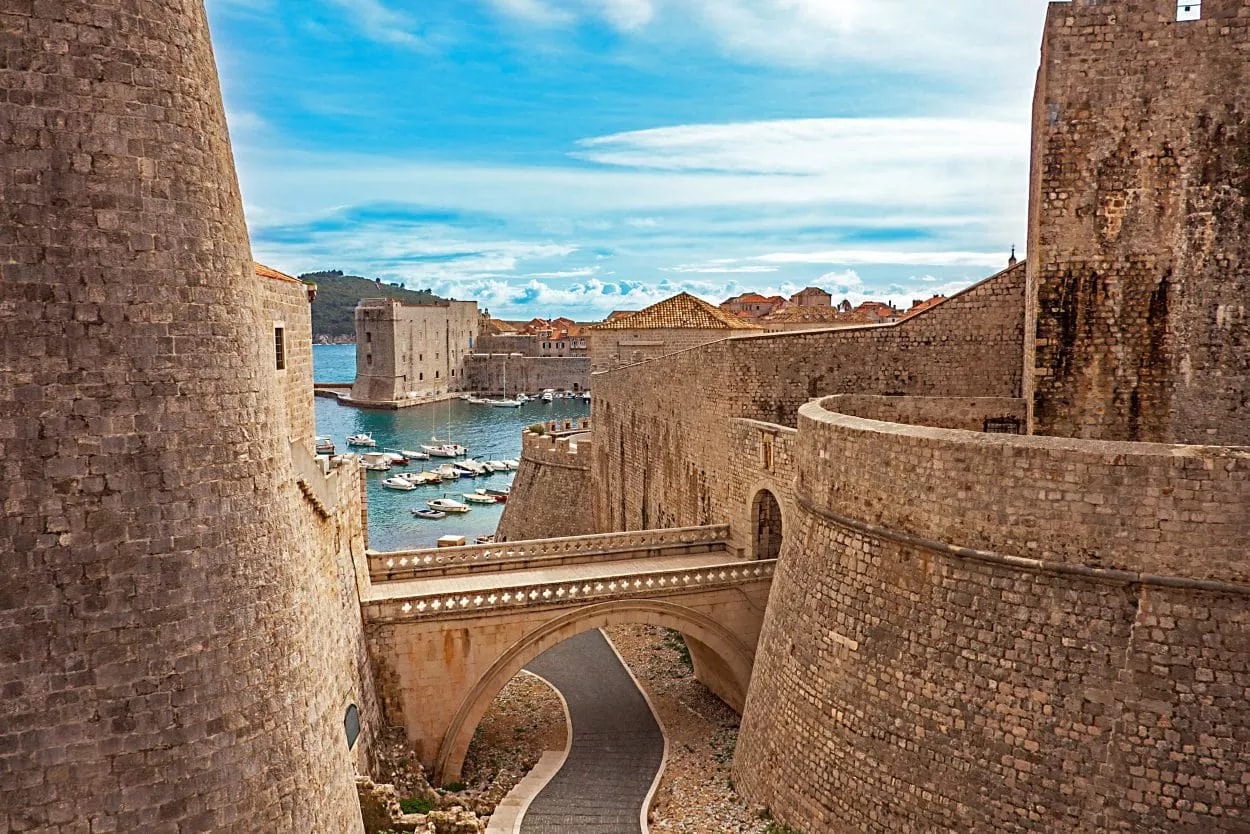Fort walls of Dubrovnik with stone bridge overlooking harbor and boats under blue sky.