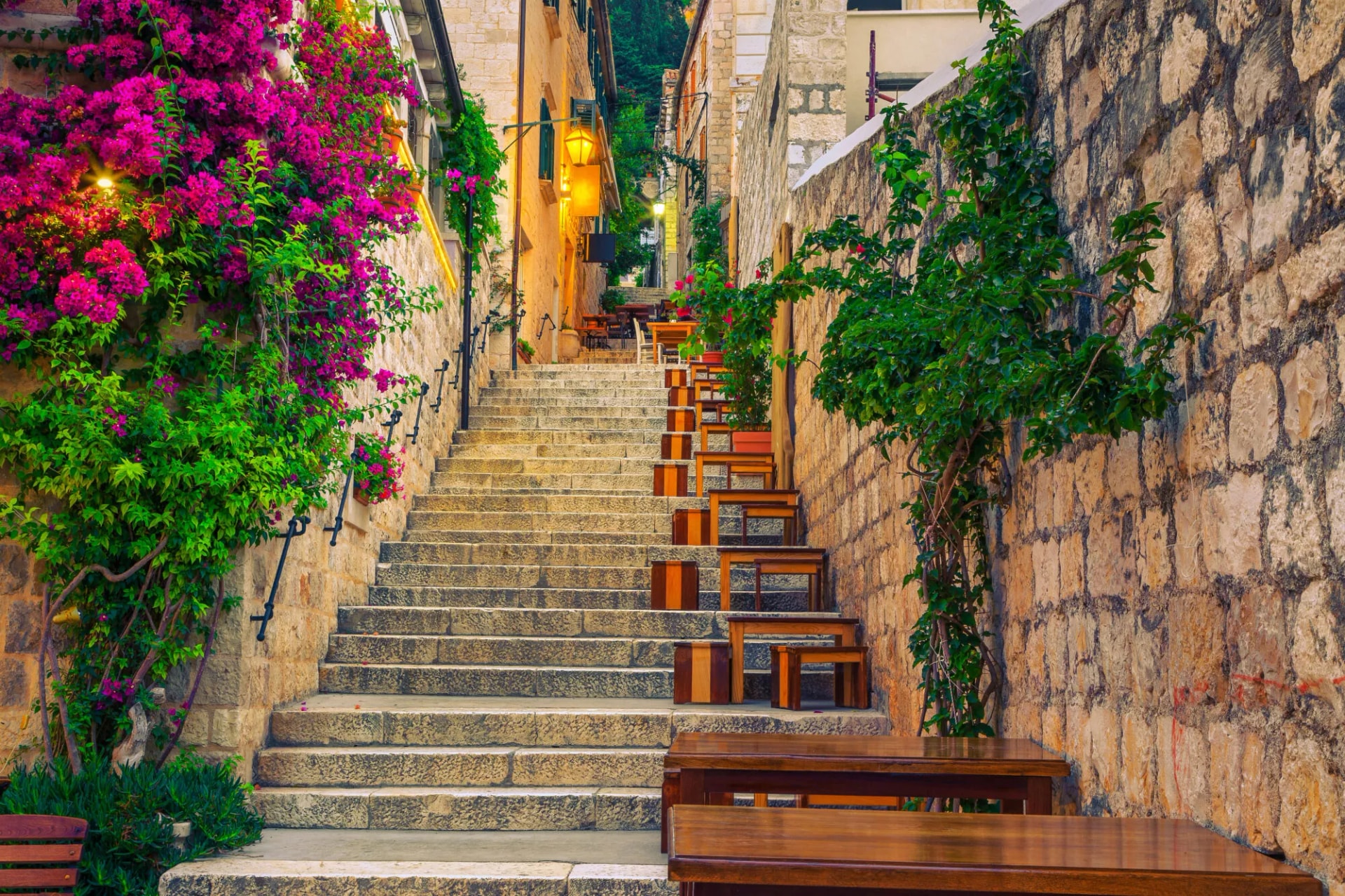 Stone staircase in a narrow alley lined with stone walls, bright pink bougainvillea, and outdoor cafe seating in Hvar.