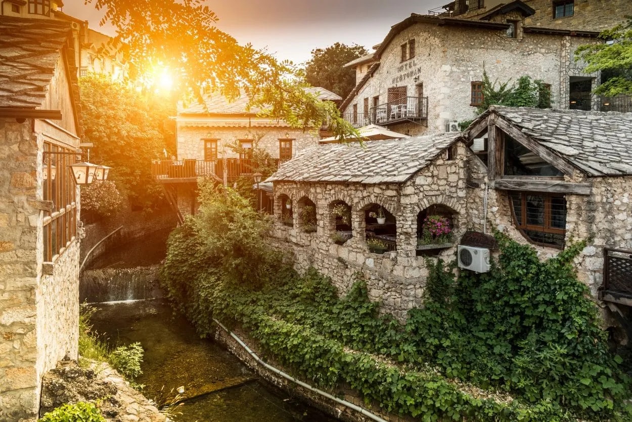 Stone buildings with slate roofs line a narrow river with a small waterfall in Mostar old town.
