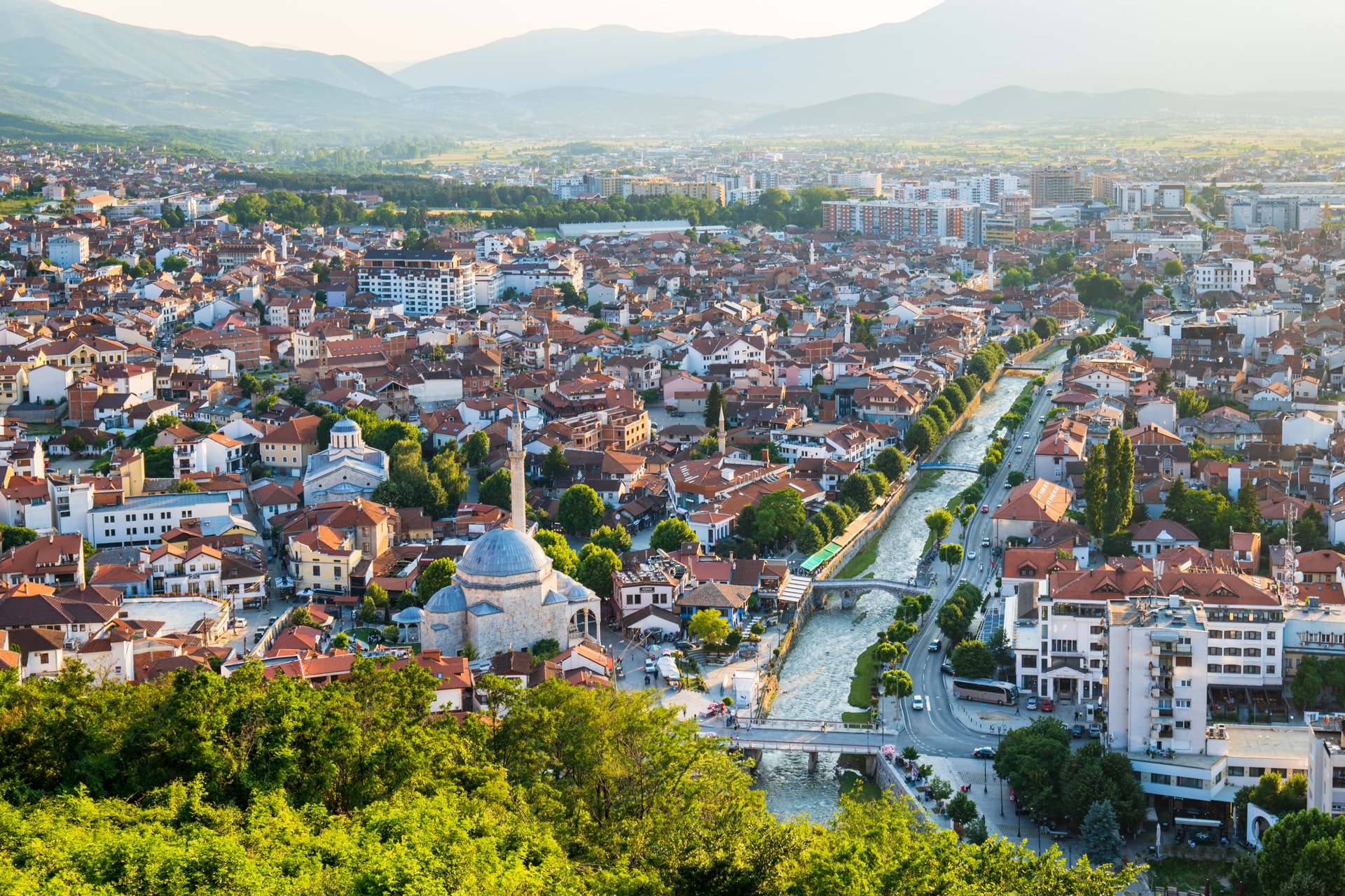 Cityscape with mosque and river flowing through dense buildings, backed by hazy mountains