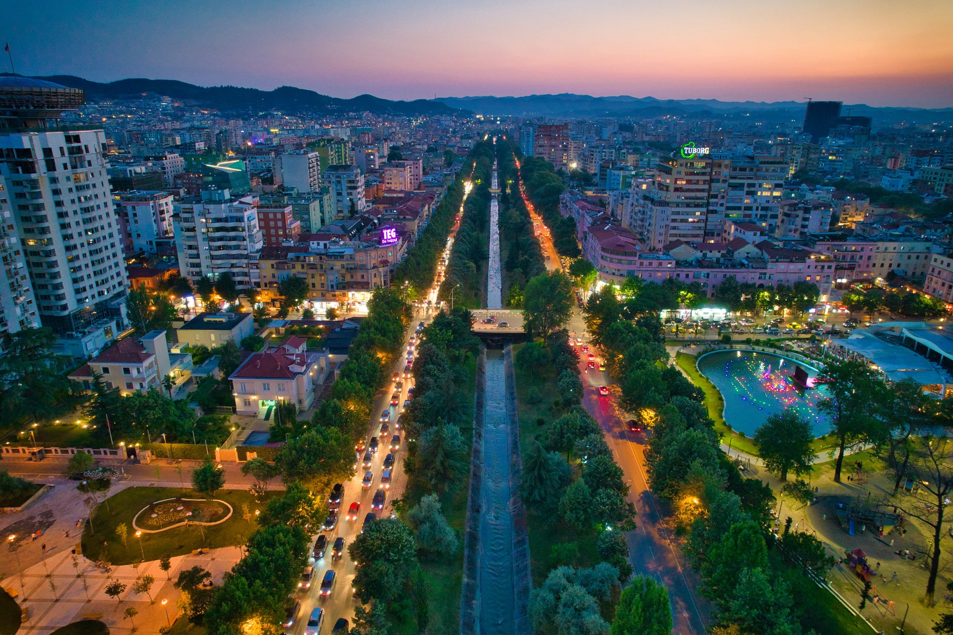 Aerial view of Tirana city center at dusk with illuminated boulevard, traffic, and mountains in background.