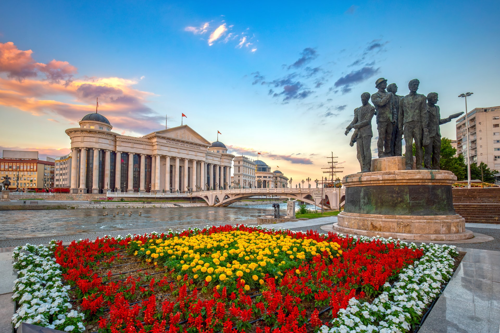 Bronze statues by river with neoclassical museum and flower beds in Skopje at sunset.