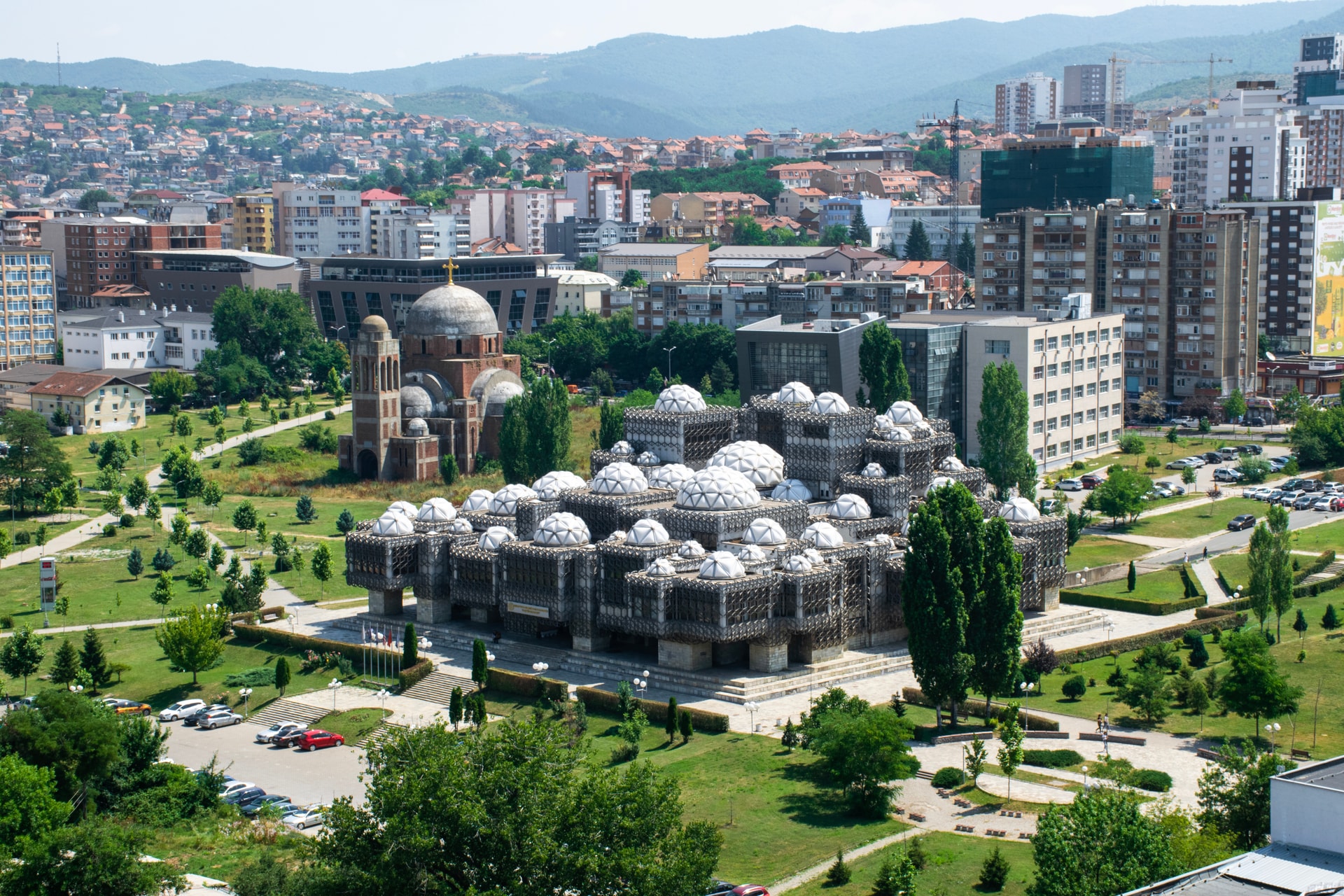 National Library of Kosovo with white domes, next to a brick church, overlooking a city and hills.