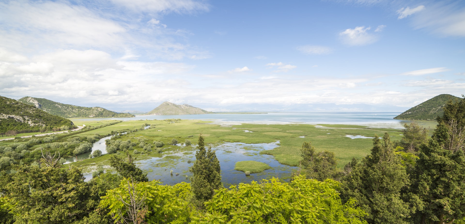 View over Lake Shkodra, Montenegro, showing marshland, green hills, and mountains under a cloudy blue sky.
