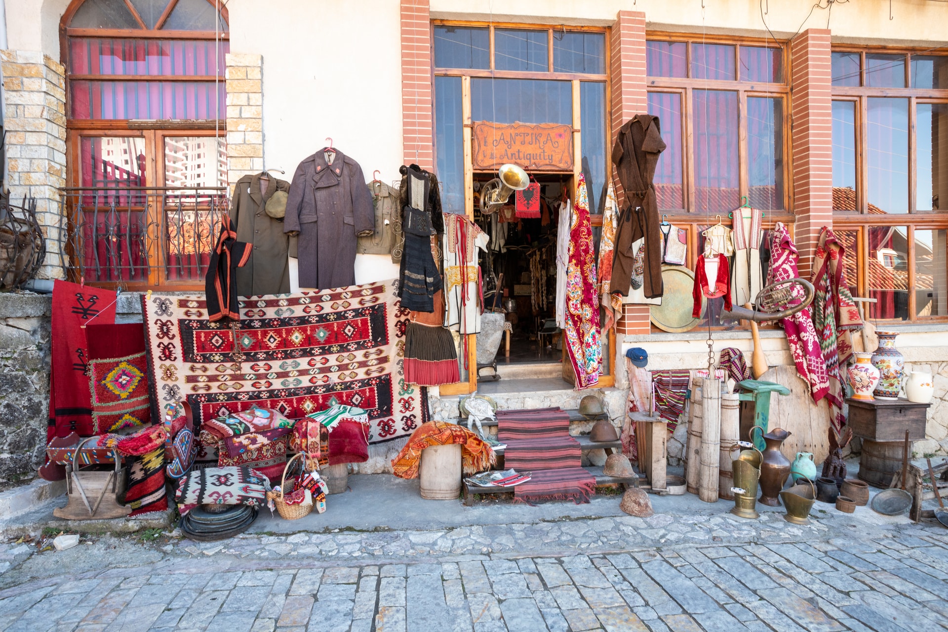 Antiquity shop exterior in Krujë with rugs, clothing, brassware, and military helmets displayed.