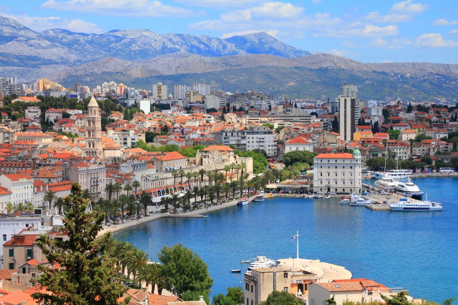 Coastal city with red roofs, bell tower, and mountains in background over blue harbor water.